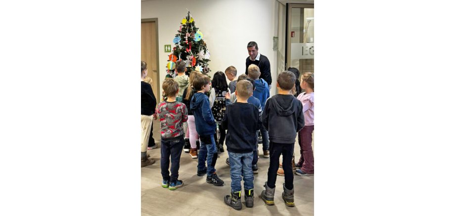 Im Foto sind die Kinder mit Bürgermeister Mark Pullmann vor dem geschmückten Weihnachtsbaum im Rathausfoyer zu sehen.